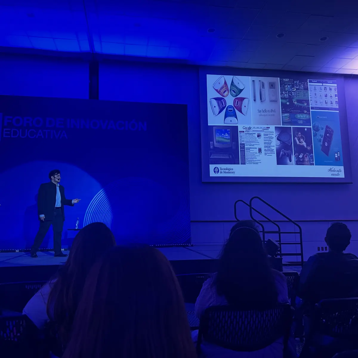 Alberto Gallegos en el escenario junto a una diapositiva con productos tecnológicos de los años 2000, como el iMac, el iPod y Facebook